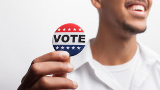 Hombre sonriente sostiene un botón rojo, blanco y azul que dice VOTE, sobre fondo blanco.