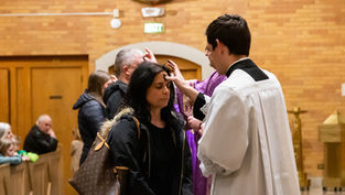 Sacerdote en sotana blanca aplica ceniza en la frente de una mujer en una iglesia de ladrillo. Fila de personas espera su turno. Atmosfera solemne.