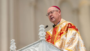 Obispo católico hablando desde un púlpito, con casulla roja y dorada, en una iglesia de fondo claro.