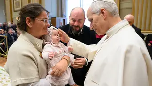 Sacerdote bendice a un bebé en brazos de una mujer. Hombre observa sonriente. Interior elegante, ambiente cálido y emotivo.