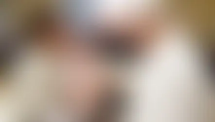 Sacerdote bendice a un bebé en brazos de una mujer. Hombre observa sonriente. Interior elegante, ambiente cálido y emotivo.