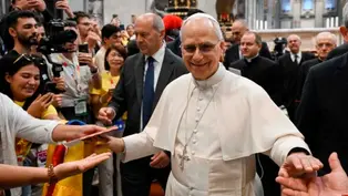 Un clérigo sonriente en túnica blanca saluda a una multitud en una iglesia grande. Personas toman fotos; ambiente alegre y colorido.
