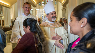 Obispo con mitra y monaguillo saludan a feligreses en una iglesia iluminada; ambiente cordial y festivo.