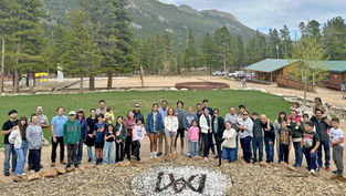 Grupo grande de familias y niños posando sonrientes en un campamento montañoso con pinos, cabañas y montañas al fondo.