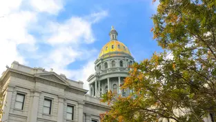 Cúpula dorada del edificio del Capitolio bajo un cielo azul con nubes. Árboles verdes y dorados en el primer plano. Ambiente sereno.