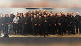 Grupo de hombres sonrientes, muchos con collares de cruces, posan en una sala. Fondo gris y suelo de alfombra. Ambiente formal.