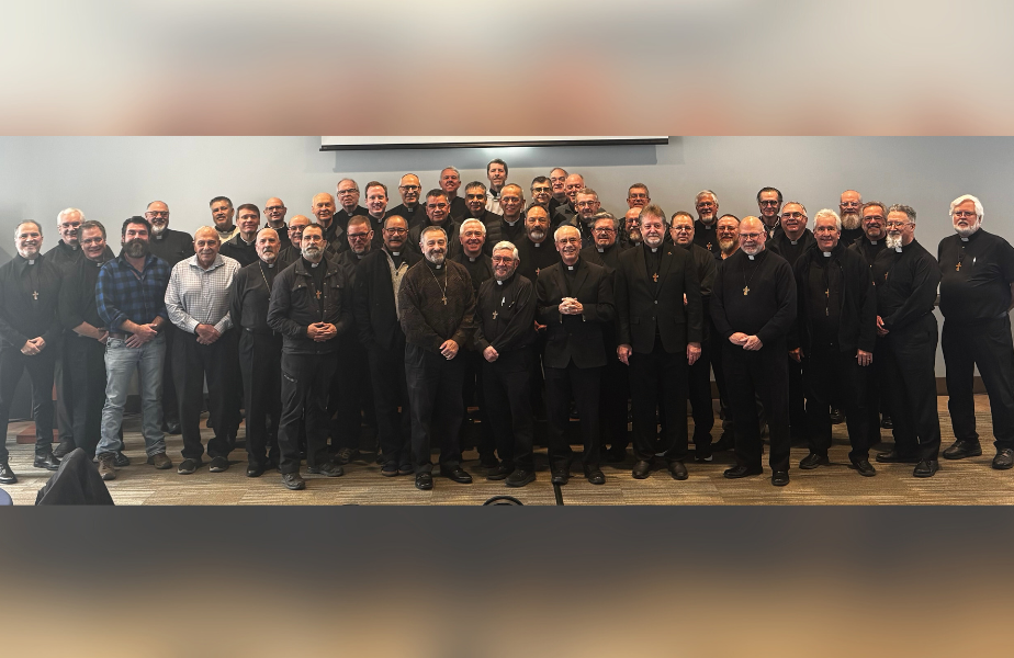 Grupo de hombres sonrientes, muchos con collares de cruces, posan en una sala. Fondo gris y suelo de alfombra. Ambiente formal.