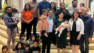 Un grupo grande de personas, incluidos adultos y niños, posan en escalones de una iglesia. Dos niños son cargados. Ambiente alegre.