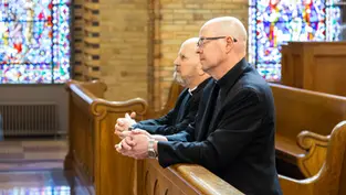 Dos hombres con vestimenta religiosa rezan en un banco de iglesia con vitrales coloridos en el fondo. Ambiente solemne.