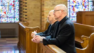 Dos hombres con vestimenta religiosa rezan en un banco de iglesia con vitrales coloridos en el fondo. Ambiente solemne.