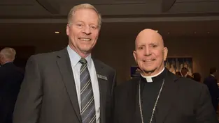 Dos hombres sonrientes en trajes formales en una sala de conferencias. Fondo oscuro con personas borrosas. Uno lleva un collar religioso.