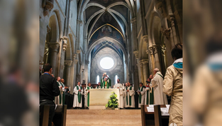Sacerdote eleva la Eucaristía en una iglesia gótica con altos techos. Varios clérigos en sotanas verdes rodean el altar. Ambiente solemne.