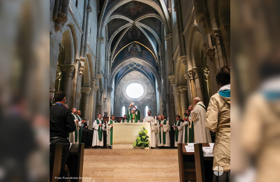 Sacerdote eleva la Eucaristía en una iglesia gótica con altos techos. Varios clérigos en sotanas verdes rodean el altar. Ambiente solemne.