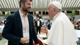 Un hombre y una figura eclesiástica conversan sonrientes en un salón amplio con techo de patrón luminoso. Ambiente cordial y formal.