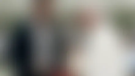 Un hombre y una figura eclesiástica conversan sonrientes en un salón amplio con techo de patrón luminoso. Ambiente cordial y formal.