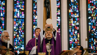 Sacerdote en misa alzando la hostia. Viste ropaje púrpura. Fondo de vidrieras coloridas. Emoción solemne.