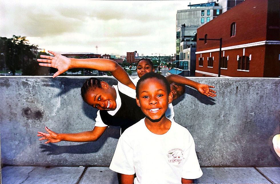 Niños sonrientes posando alegremente en una terraza urbana; edificio rojo al fondo. Ropa blanca destaca sobre el muro gris. Cielo nublado.