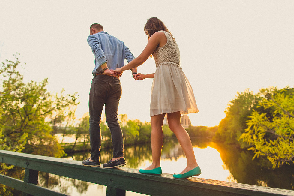 Pareja caminando de la mano sobre una baranda, rodeados de árboles al atardecer. Ambiente romántico con luz cálida y reflejos en el agua.