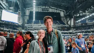 Dos personas sonríen en un estadio lleno de gente con luces brillantes. Visten de manera casual y llevan gafetes. Ambiente alegre y concurrido.