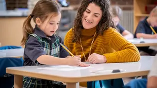 Niña y mujer en aula, la niña escribe mientras la mujer en suéter amarillo sonríe. Fondo desenfocado, ambiente educativo y cálido.
