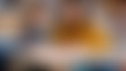 Niña y mujer en aula, la niña escribe mientras la mujer en suéter amarillo sonríe. Fondo desenfocado, ambiente educativo y cálido.