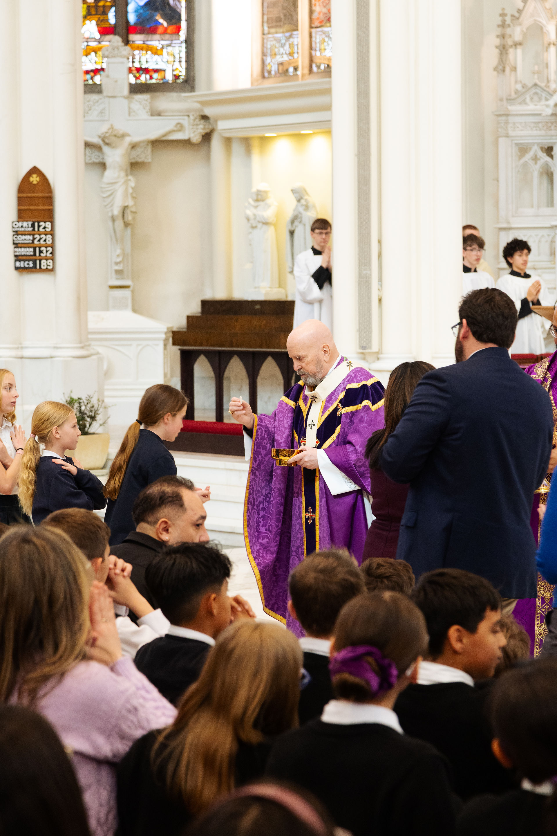 Obispo en túnica morada da la comunión a niños en una iglesia con vitrales. Ambiente solemne, asistentes observan respetuosamente.