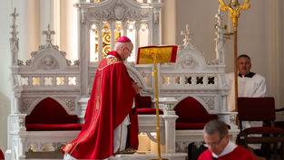 Obispo católico de rojo orando ante un atril en una iglesia ornamentada, con ayudantes al fondo y ambiente solemne.