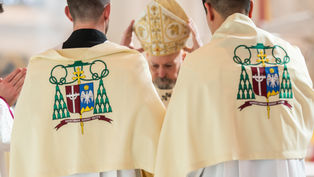 Dos personas con capas bordadas, de espaldas, frente a un hombre con mitra dorada levantando las manos. Interior de iglesia. Ambiente solemne.