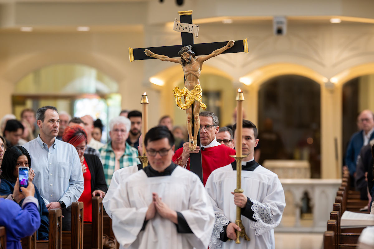 Una procesión avanza por la catedral con un crucifijo elevado, acompañado por cirios y monaguillos.