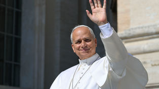 El papa León, con sotana blanca, saluda con la mano desde un balcón; fondo de piedra y luz soleada.