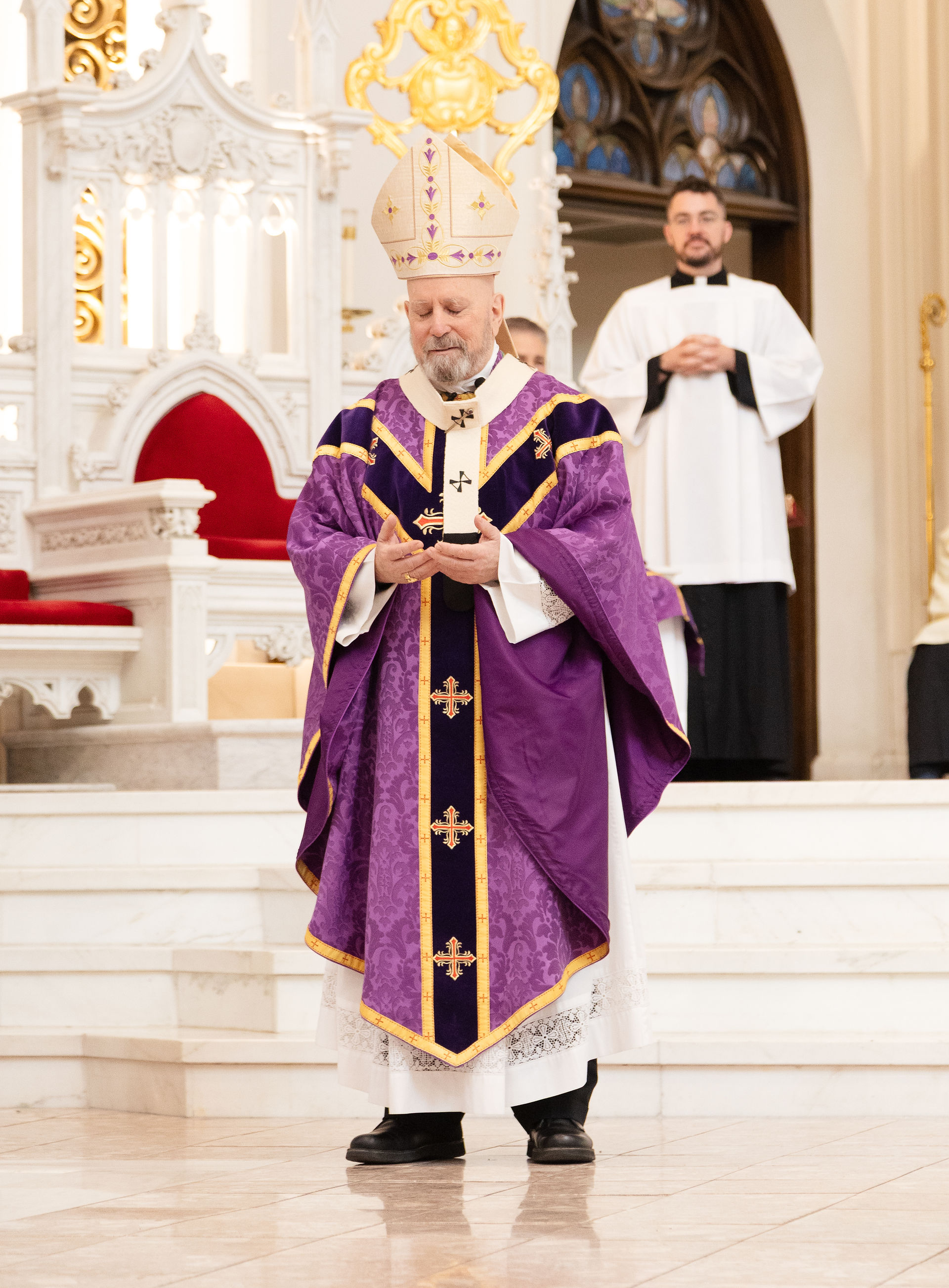 Sacerdote con traje púrpura en misa, sosteniendo un objeto. Fondo de catedral con púlpito blanco y dorado. Hombre de pie detrás. Ambiente solemne.