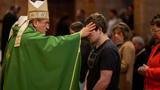 Obispo con vestimenta verde bendice a un joven en iglesia. Otros feligreses observan. Ambiente solemne y espiritual.
