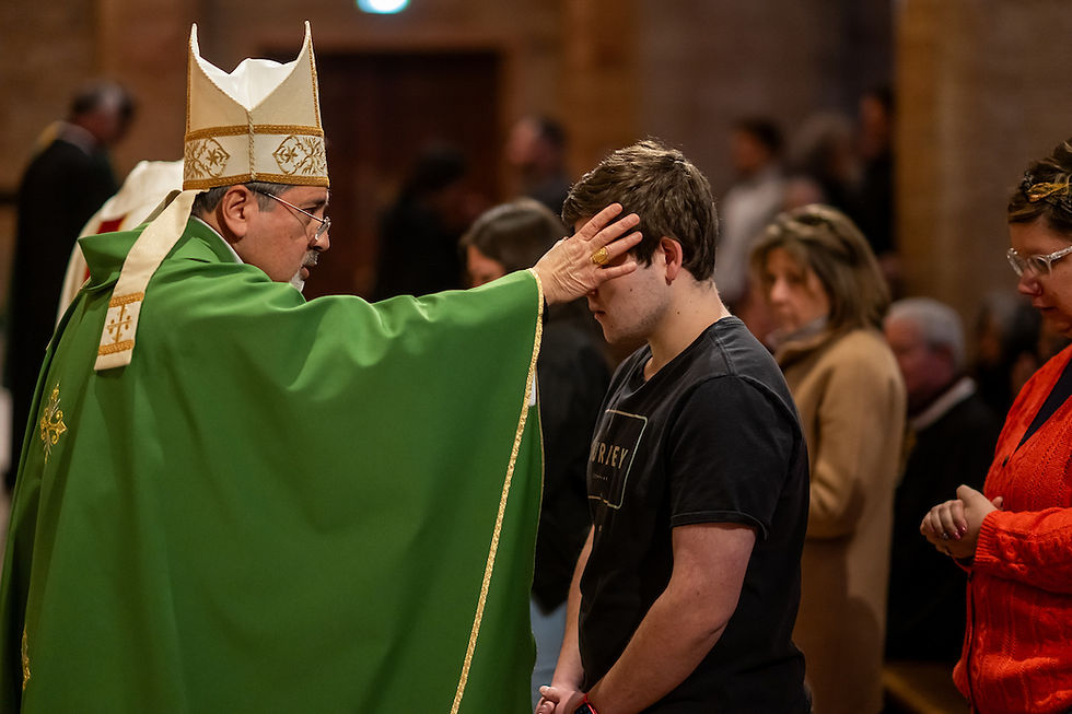 Obispo con vestimenta verde bendice a un joven en iglesia. Otros feligreses observan. Ambiente solemne y espiritual.