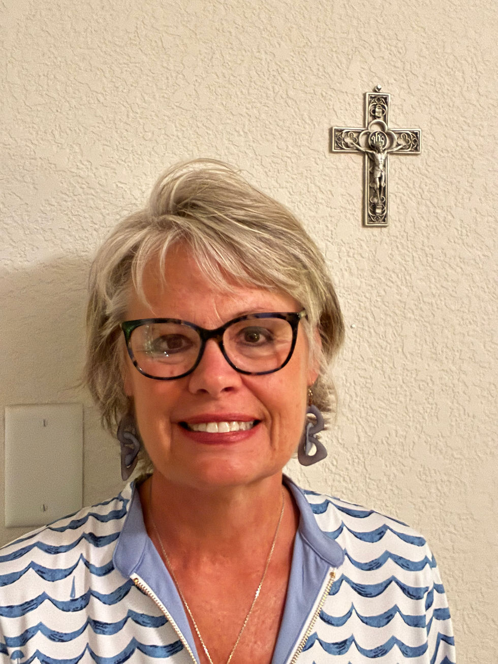 Mujer sonriente con gafas y cabello gris, frente a una pared beige con un crucifijo; lleva blusa azul y blanca a rayas.