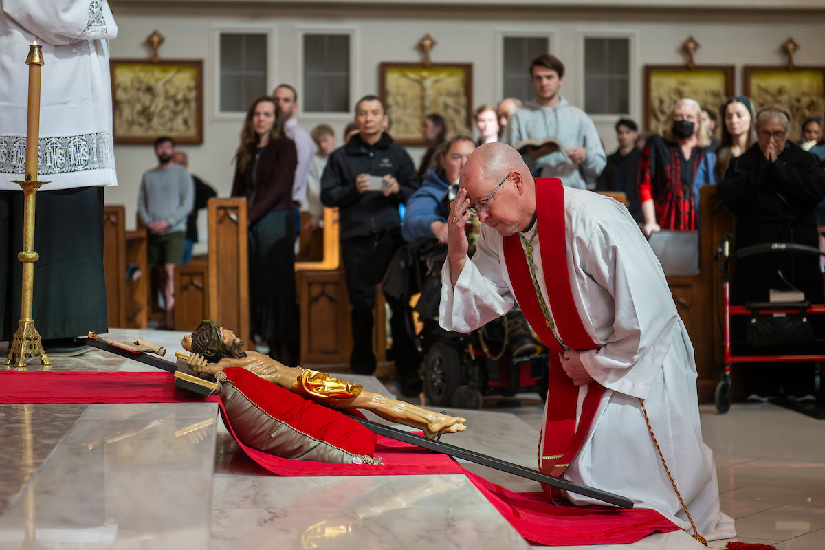 Un sacerdote se arrodilla y venera la cruz, inclinando la cabeza ante un crucifijo sobre una tela roja mientras la congregación observa.