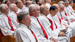 Hombres mayores con túnicas blancas y estolas rojas, sentados en silencio en una iglesia, con gesto solemne.