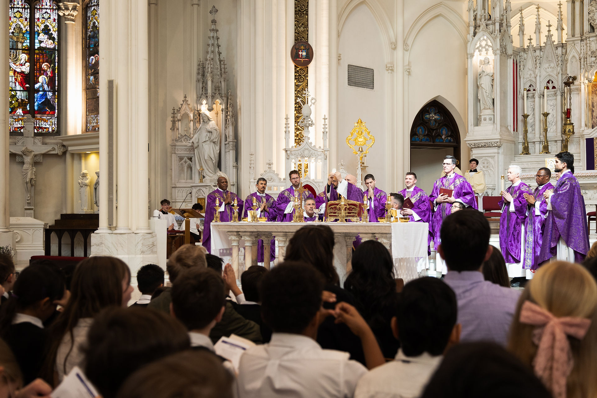 Sacerdotes en túnicas moradas celebran misa en una iglesia gótica llena de gente; vitrales coloridos al fondo, ambiente solemne.
