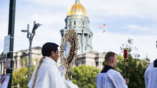 Procesión católica al aire libre: sacerdotes con una custodia dorada ante una cúpula dorada; cielo despejado y solemne.