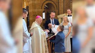 Ceremonia de bautizo en iglesia. Un sacerdote con mitra, padres y niño con bebé en brazos. Ambiente solemne, interior de ladrillo.