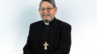 Hombre mayor con gafas y barba, vestido de sacerdote con cruz dorada. Fondo blanco. Expresión amable. Traje negro formal.
