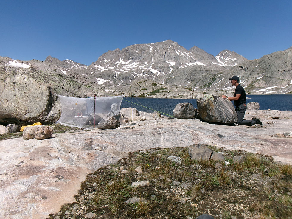 Campista arrodillado junto a una roca en un campamento alpino, con tienda mosquitera, lago azul y montañas nevadas bajo cielo despejado.