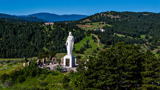 Estatua blanca de Jesús en un mirador montañoso, rodeada de pinos y colinas verdes bajo cielo azul despejado.