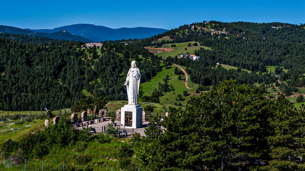 Estatua blanca de Jesús en un mirador montañoso, rodeada de pinos y colinas verdes bajo cielo azul despejado.