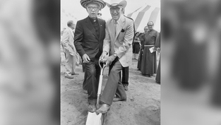 Dos hombres con sombrero posan sobre una pala en una ceremonia de inauguración, con carpas y clérigos detrás.
