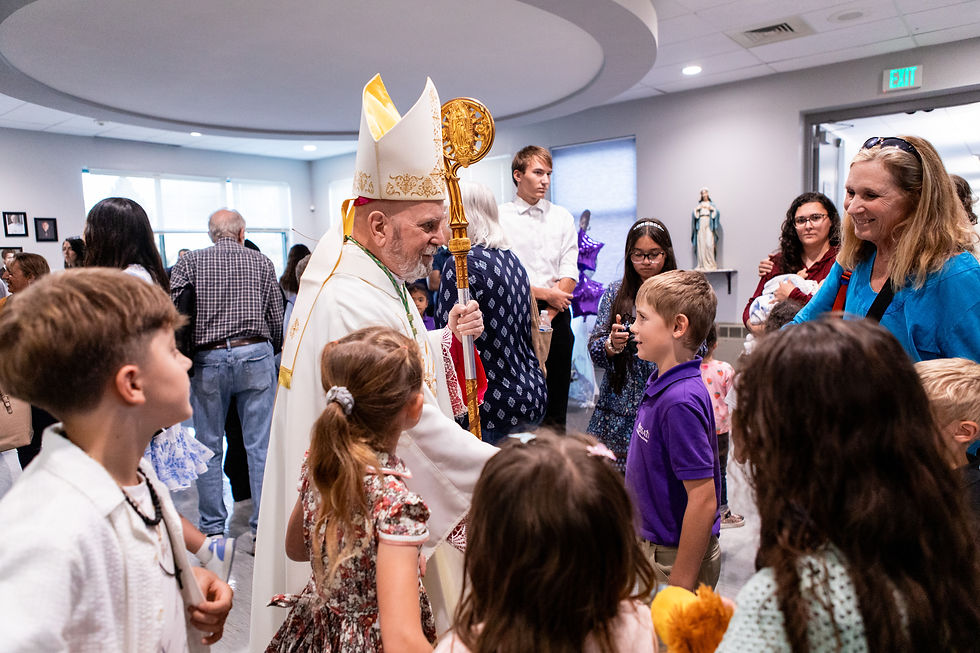 Un sacerdote con mitra y báculo saluda a un grupo de niños en una sala iluminada, rodeado de adultos. Ambiente festivo y alegre.