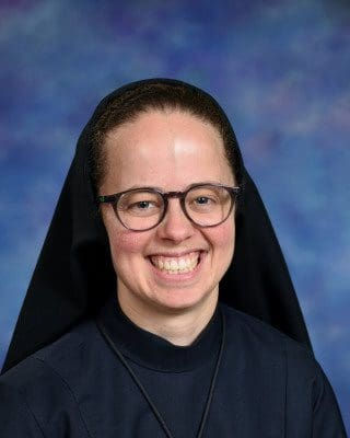 Retrato de una monja sonriente con gafas y hábito oscuro, sobre fondo azul difuminado.
