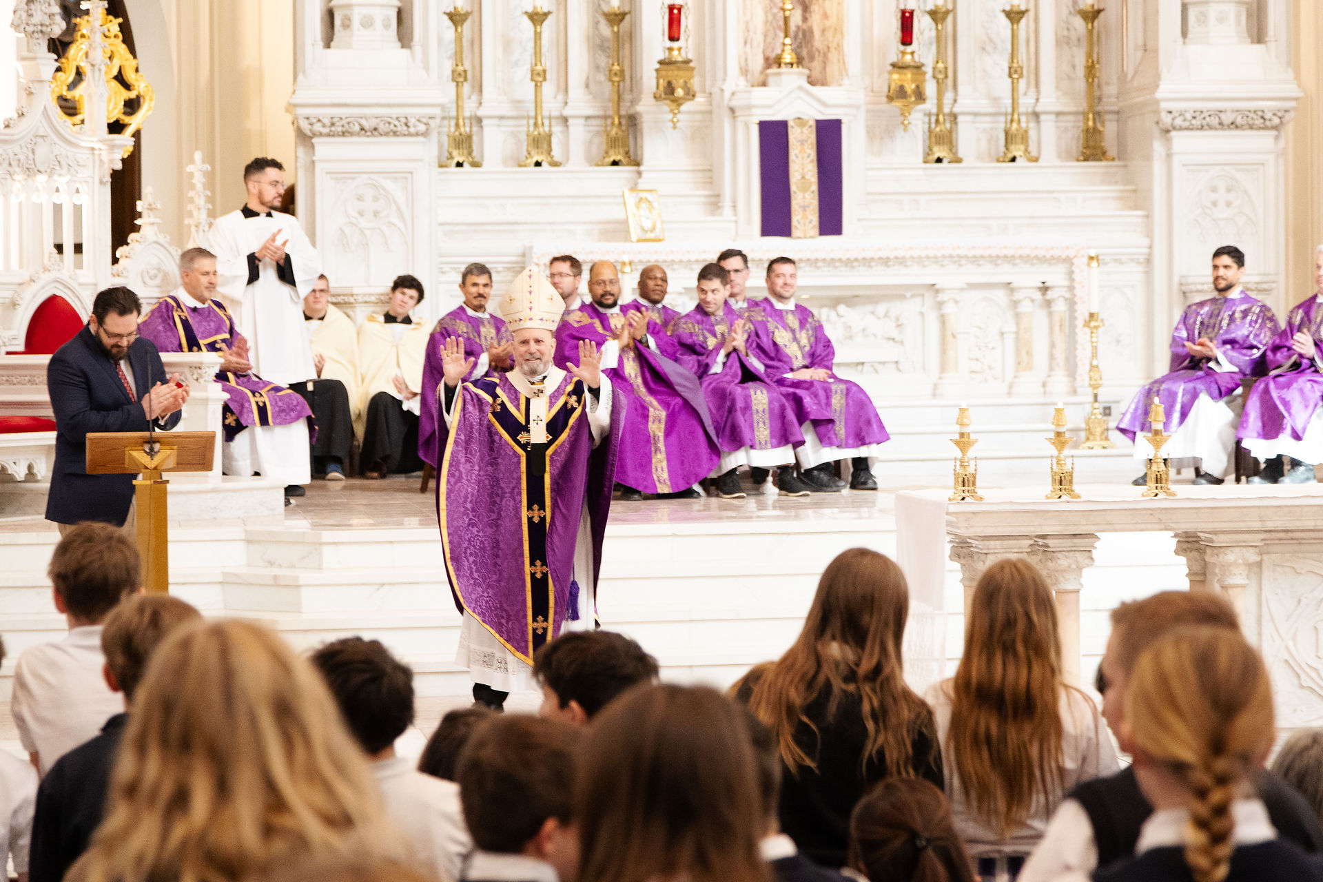 Obispo con vestiduras moradas se dirige a un grupo de estudiantes frente al altar, mientras varios sacerdotes están sentados detrás de él en el santuario de la iglesia.