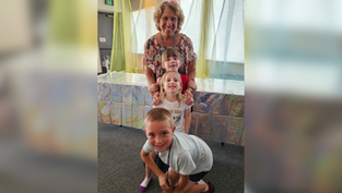 Mujer sonriente con tres niños en sala decorada con telas de colores y mesa brillante; ambiente alegre.