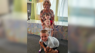 Mujer sonriente con tres niños en sala decorada con telas de colores y mesa brillante; ambiente alegre.