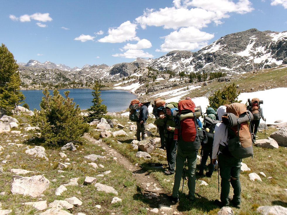 Grupo de senderistas con mochilas avanza junto a un lago alpino, entre rocas y nieve, bajo cielo azul soleado.